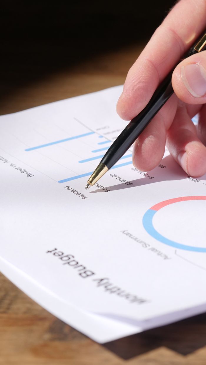who-we-are Close-up of hand analyzing data on a monthly budget document with graphs and charts.